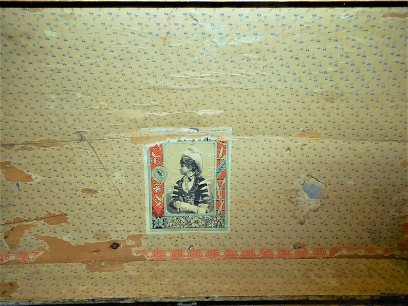 May include: A vintage suitcase with a faded floral pattern and a small, rectangular label featuring a woman in a hat. The label has a blue and red border and a floral design.