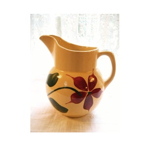 May include: A cream-colored ceramic pitcher with a green and red floral design. The pitcher has a round body and a handle.