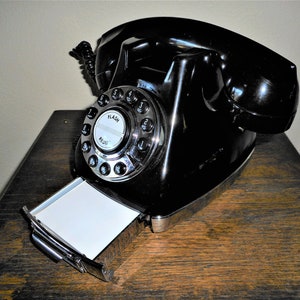 K&ouml;nnte beinhalten: Ein schwarzes W&auml;hlscheiben-Telefon mit einem verchromten W&auml;hlrad und einem herausziehbaren Notizblock. Das Telefon steht auf einer Holzoberfl&auml;che.