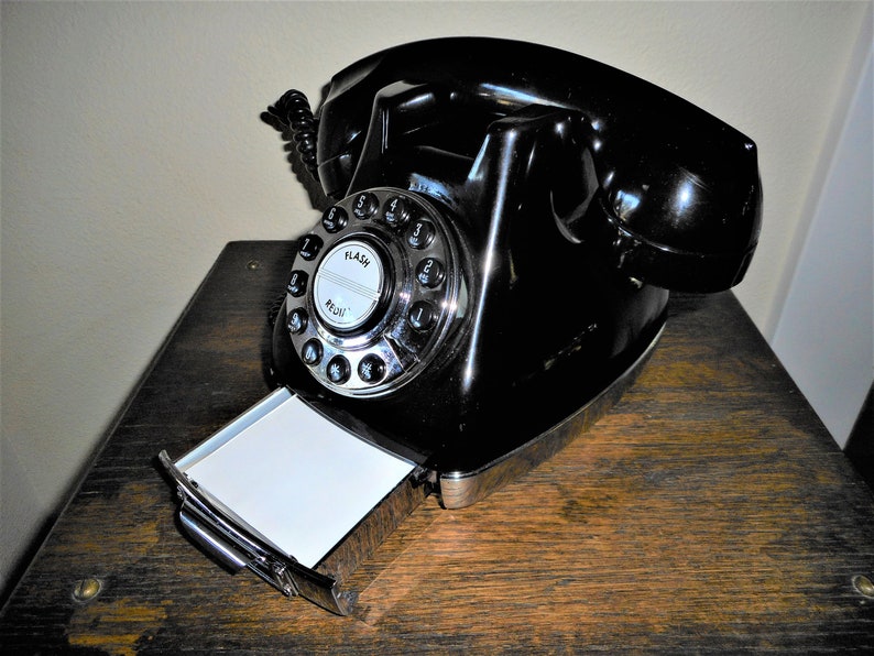 K&ouml;nnte beinhalten: Ein schwarzes W&auml;hlscheiben-Telefon mit einem verchromten W&auml;hlrad und einem herausziehbaren Notizblock. Das Telefon steht auf einer Holzoberfl&auml;che.