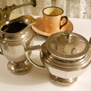 May include: A silver-colored tea set with a creamer and a lidded sugar bowl. A peach-colored cup with white polka dots and a matching saucer are also present. The creamer is filled with a dark liquid. A decorative brass object is in the background.