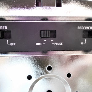 K&ouml;nnte beinhalten: Nahaufnahme eines silbernen Telefons mit schwarzen Kn&ouml;pfen und Beschriftungen. Die Beschriftungen lauten "RINGER", "ON", "OFF", "TONE", "PULSE", "RECEIVER VOLUME", "HI" und "LOW".