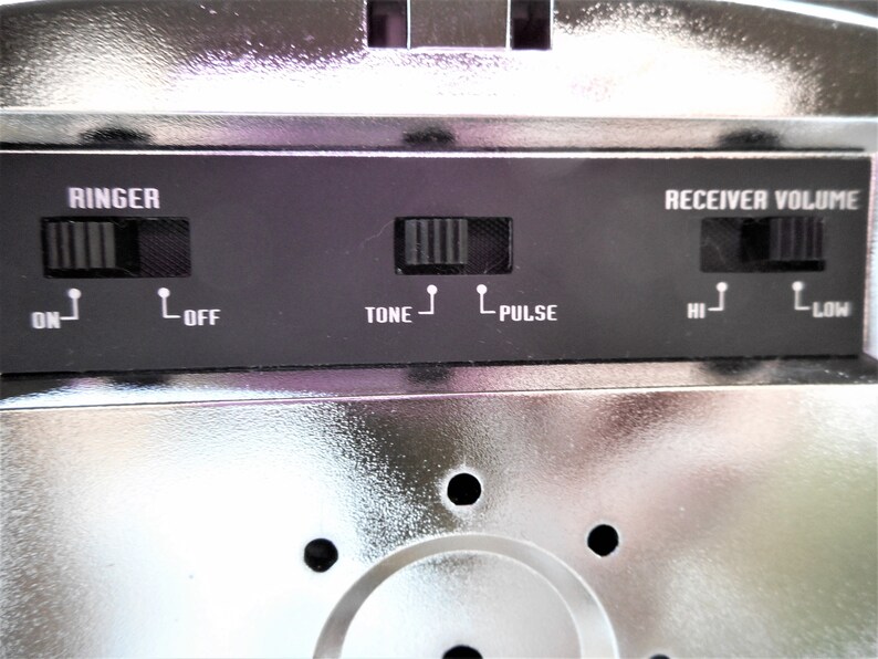 K&ouml;nnte beinhalten: Nahaufnahme eines silbernen Telefons mit schwarzen Kn&ouml;pfen und Beschriftungen. Die Beschriftungen lauten "RINGER", "ON", "OFF", "TONE", "PULSE", "RECEIVER VOLUME", "HI" und "LOW".