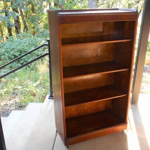 May include: A tall, wooden bookcase with four shelves. The bookcase is dark brown and has a simple design. It is standing on a concrete surface.