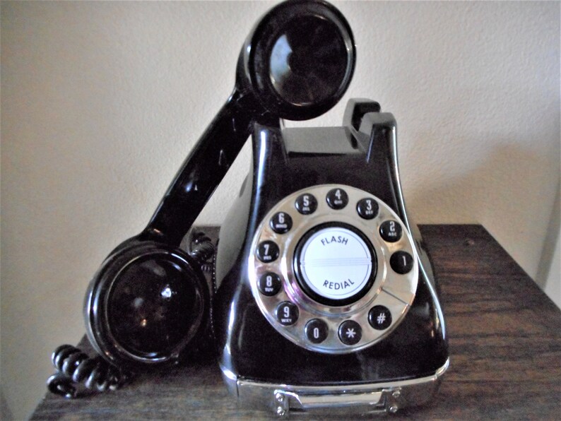 K&ouml;nnte beinhalten: Ein schwarzes W&auml;hlscheibentelefon mit silbernem Frontblech und Kn&ouml;pfen. Das Telefon hat ein schwarzes H&ouml;rer mit einem schwarzen Kabel. Das Telefon hat die W&ouml;rter "FLASH" und "REDIAL" auf dem Frontblech aufgedruckt.