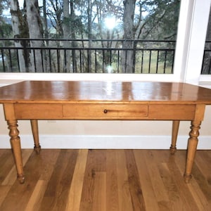 May include: A light brown wooden table with a single drawer and turned legs. The table has a rectangular top with rounded corners. The table is set on a hardwood floor in front of a window.