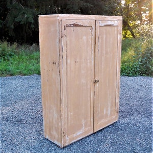 May include: A tall, two-door cabinet made of wood with a distressed, light brown paint finish. The cabinet has simple, flat doors with no hardware visible.