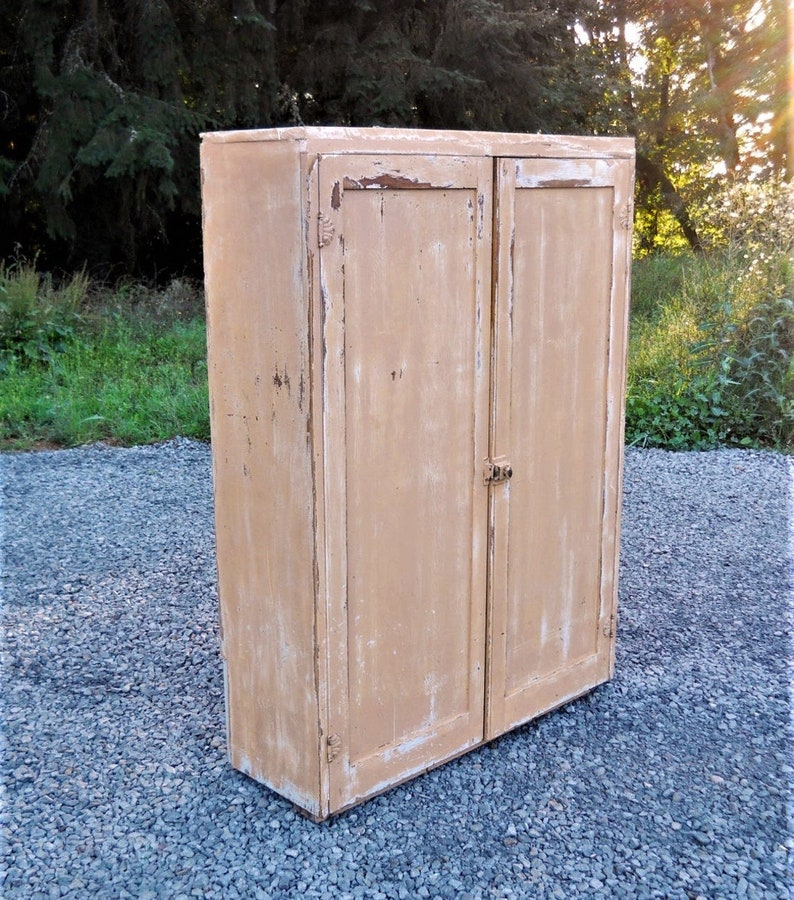 May include: A tall, two-door cabinet made of wood with a distressed, light brown paint finish. The cabinet has simple, flat doors with no hardware visible.