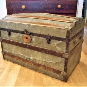 May include: A vintage, weathered, gray metal and wood travel trunk with metal hardware and a wooden latch. The trunk has a rounded top and is sitting on a wooden floor.