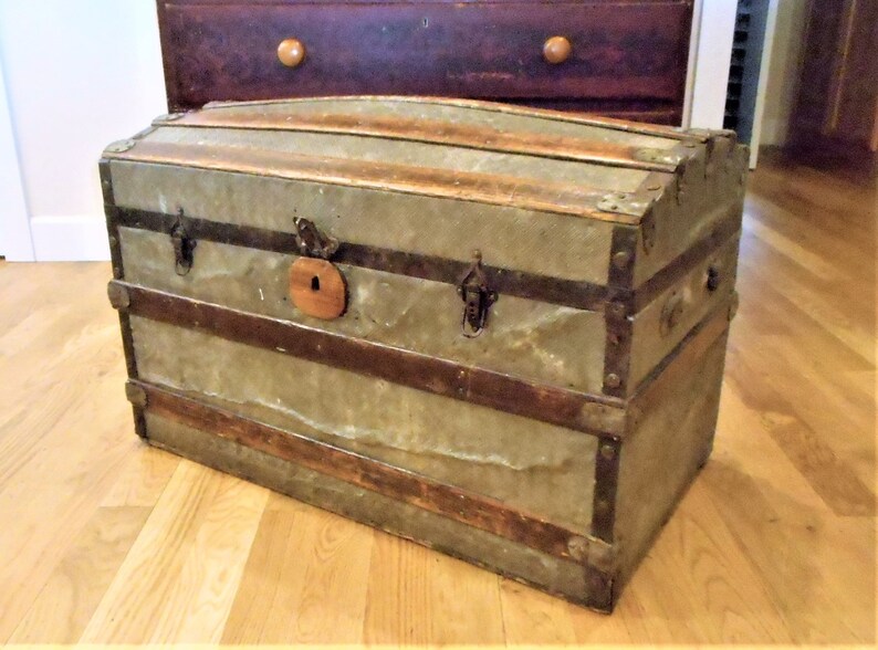 May include: A vintage, weathered, gray metal and wood travel trunk with metal hardware and a wooden latch. The trunk has a rounded top and is sitting on a wooden floor.