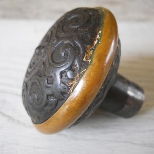 May include: A black door knob with a decorative, ornate design and a gold-colored rim. The knob has a black metal stem.