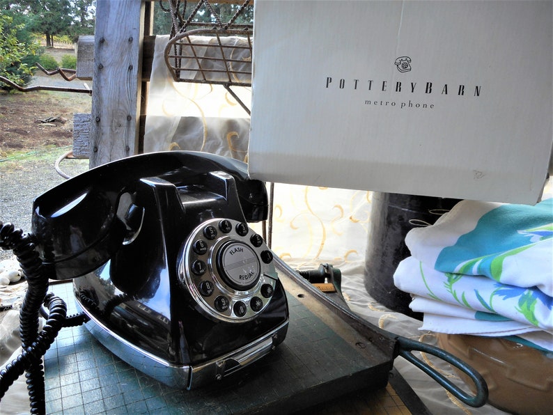 K&ouml;nnte beinhalten: Ein schwarzes W&auml;hlscheibentelefon mit silberner Frontplatte und wei&szlig;em Zifferblatt. Das Telefon hat ein schwarzes Kabel und steht auf einer gr&uuml;nen Oberfl&auml;che. Das Telefon ist mit "FLASH" und "PULSE" beschriftet.