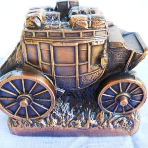 May include: A bronze-coloured metal money box shaped like a stagecoach with a detailed design. The stagecoach has a wooden frame and is filled with luggage. The stagecoach has two wheels and is sitting on a base of grass.