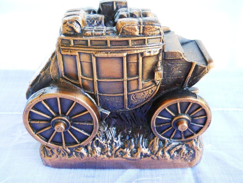 May include: A bronze-coloured metal money box shaped like a stagecoach with a detailed design. The stagecoach has a wooden frame and is filled with luggage. The stagecoach has two wheels and is sitting on a base of grass.