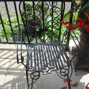 May include: A black metal chair with a decorative, ornate design. The chair has a slatted seat and a high back. The chair is sitting on a concrete patio.