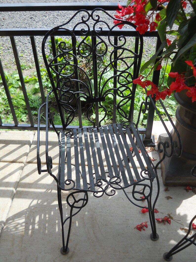 May include: A black metal chair with a decorative, ornate design. The chair has a slatted seat and a high back. The chair is sitting on a concrete patio.