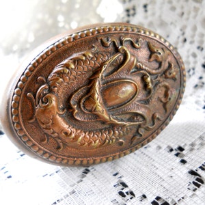 May include: An oval-shaped, antique, copper-colored trinket box with a raised, embossed design of two fish with a crown above them.