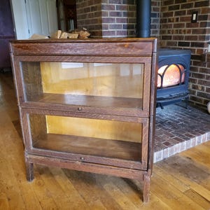 May include: A two-shelf wooden bookcase with glass doors. The bookcase is made of dark wood and has a simple design. It is standing on a wooden floor in front of a brick fireplace.