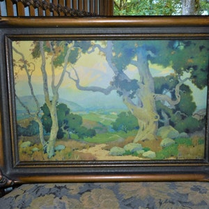 May include: A framed landscape painting depicting a scenic view of a valley with trees and hills. The painting is done in a soft, muted palette of greens, blues, and yellows. The frame is a dark wood with a gold-colored trim.