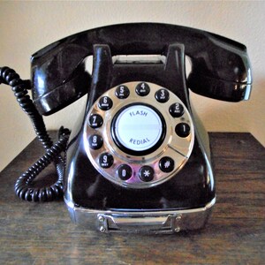 K&ouml;nnte beinhalten: Ein schwarzes W&auml;hlscheiben-Telefon mit einer silbernen Frontplatte und einem wei&szlig;en Zifferblatt. Das Zifferblatt hat die Zahlen 1 bis 9, 0, *, und #. Das Telefon hat einen silbernen Knopf mit der Aufschrift "FLASH" und "REDIAL".