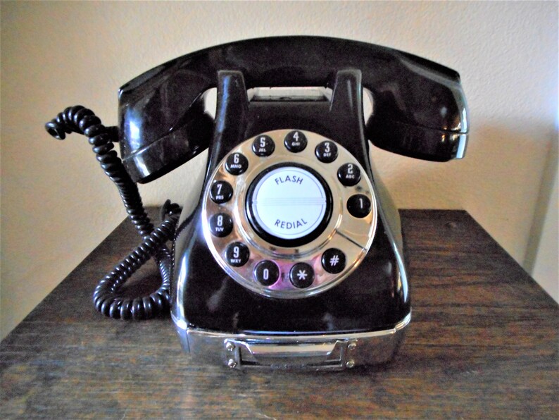 K&ouml;nnte beinhalten: Ein schwarzes W&auml;hlscheiben-Telefon mit einer silbernen Frontplatte und einem wei&szlig;en Zifferblatt. Das Zifferblatt hat die Zahlen 1 bis 9, 0, *, und #. Das Telefon hat einen silbernen Knopf mit der Aufschrift "FLASH" und "REDIAL".