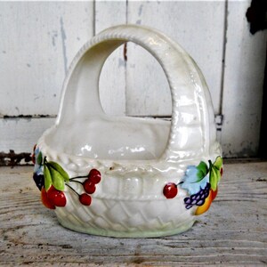 May include: White ceramic basket with a handle, decorated with painted fruit and leaves. The basket is shaped like a woven basket with a white background and colourful fruit and leaves.