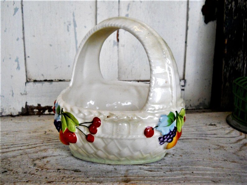 May include: White ceramic basket with a handle, decorated with painted fruit and leaves. The basket is shaped like a woven basket with a white background and colourful fruit and leaves.