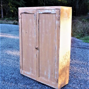May include: A vintage, two-door cabinet with a distressed, light brown paint finish. The cabinet has a simple design with a flat top and a slightly angled base.