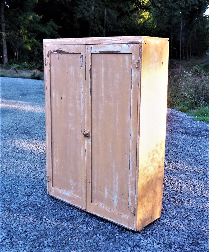 May include: A vintage, two-door cabinet with a distressed, light brown paint finish. The cabinet has a simple design with a flat top and a slightly angled base.