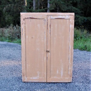 May include: A vintage wooden cabinet with two doors painted in a light peach color. The cabinet has a distressed finish with visible wood grain and some paint chipping. The doors have simple metal hinges and a small metal latch.