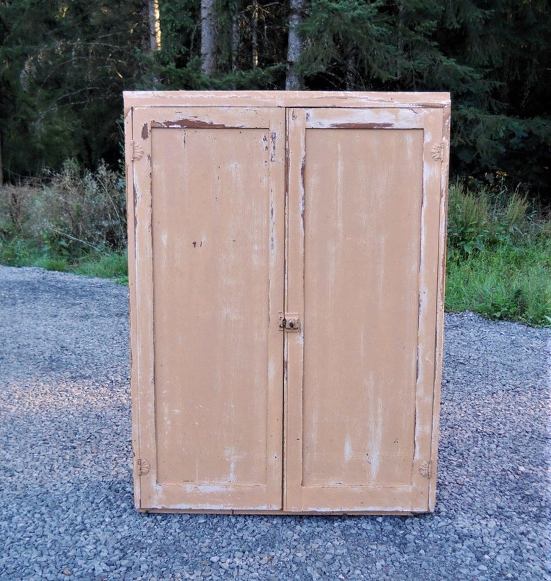 May include: A vintage wooden cabinet with two doors painted in a light peach color. The cabinet has a distressed finish with visible wood grain and some paint chipping. The doors have simple metal hinges and a small metal latch.