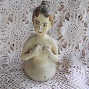 May include: A vintage ceramic doll with painted features, wearing a simple white dress. The doll has a painted red mouth and a white nose. The doll is standing on a white crocheted surface.