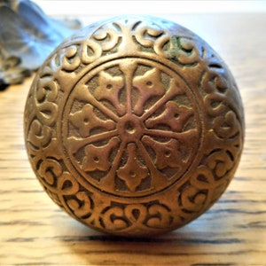May include: A round, brass door knob with an intricate, embossed design. The knob features a central circular motif with radiating lines and a surrounding border of repeating patterns.