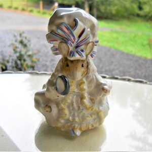 May include: A porcelain figurine of a mouse wearing a white bonnet with red, white, and blue ribbons. The mouse is holding a small, round mirror and is dressed in a white dress with yellow and blue floral details.