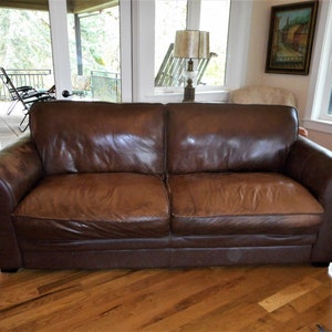 May include: A brown leather sofa with a slightly worn look. The sofa has a tufted back and rolled arms. It is sitting on a hardwood floor.
