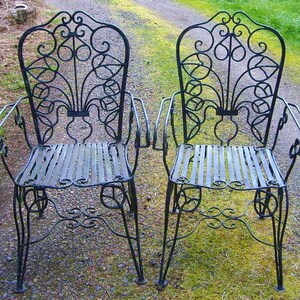 May include: Two black wrought iron chairs with intricate scrollwork designs. The chairs have a slatted seat and back.