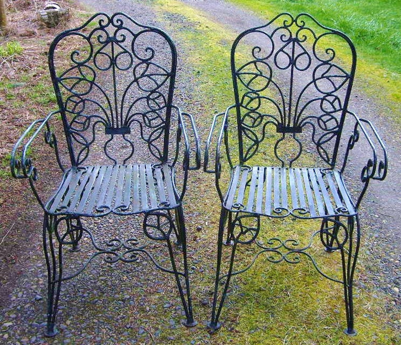 May include: Two black wrought iron chairs with intricate scrollwork designs. The chairs have a slatted seat and back.