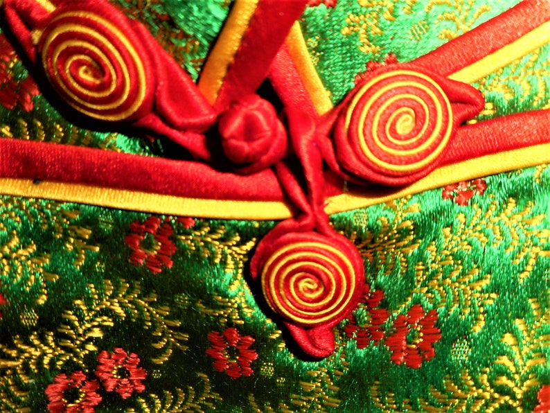 K&ouml;nnte beinhalten: Nahaufnahme eines gr&uuml;nen Stoffes mit einem goldenen und roten Blumenmuster. Der Stoff hat rote und goldene Kn&ouml;pfe mit einem Spiralmuster.