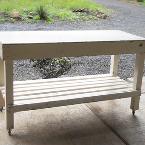 May include: White wooden table with a shelf and wheels. The table has a distressed white paint finish and is made of wood planks.