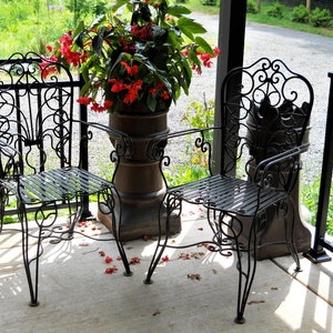 May include: Two black wrought iron chairs with ornate designs sit on a porch with a potted plant in the background. The chairs have a slatted seat and a decorative back.