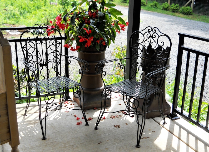 May include: Two black wrought iron chairs with ornate designs sit on a porch with a potted plant in the background. The chairs have a slatted seat and a decorative back.