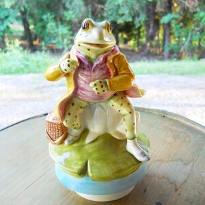 May include: A whimsical ceramic frog figurine, dressed in a yellow coat and pink vest, holding a basket. The frog is perched on a lily pad base with a light blue rim. The frog's legs are spotted, and it has a charming, expressive face.