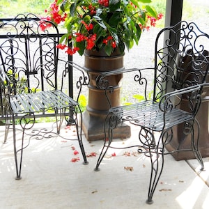 May include: Two black wrought iron chairs with ornate designs on the backrests and seats. The chairs are on a concrete patio with red flower petals scattered on the ground.