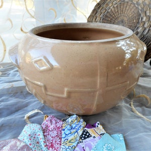 May include: A large, brown ceramic crock with a geometric design. The crock has a wide opening and a rounded body. It is sitting on a white tablecloth with a few colorful fabric scraps.