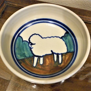 May include: A white ceramic bowl with a blue and green rim. The bowl features a hand-painted design of a white sheep in a field.