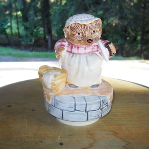 May include: A ceramic musical figurine of a hedgehog wearing a red and white checkered dress and a white bonnet. The hedgehog is holding a basket of bread and a rolling pin. The figurine is standing on a stone base.