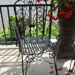 May include: A black wrought iron chair with a decorative scroll design on the back and seat. The chair is sitting on a concrete patio with red flowers in the background.