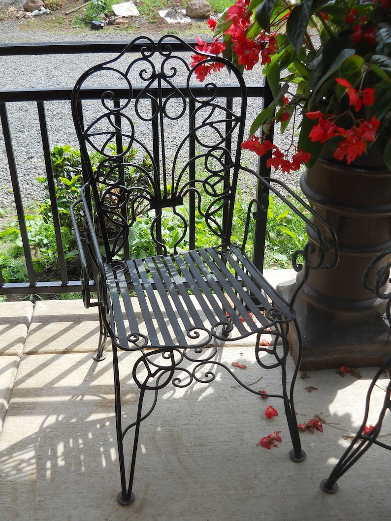 May include: A black wrought iron chair with a decorative scroll design on the back and seat. The chair is sitting on a concrete patio with red flowers in the background.