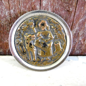 May include: A silver and brass circular pin with a raised design of a woman and a wolf in a forest setting.