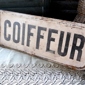 Könnte beinhalten: Ein weißes Metallschild mit schwarzer Beschriftung, auf dem "COIFFEUR" steht. Das Schild hat einen rustikalen, Vintage-Look und ist leicht verwittert.